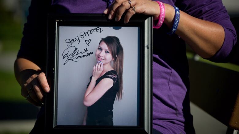 A woman holds a framed photo of a teenaged girl with long brown hair wearing a black dress. The words 