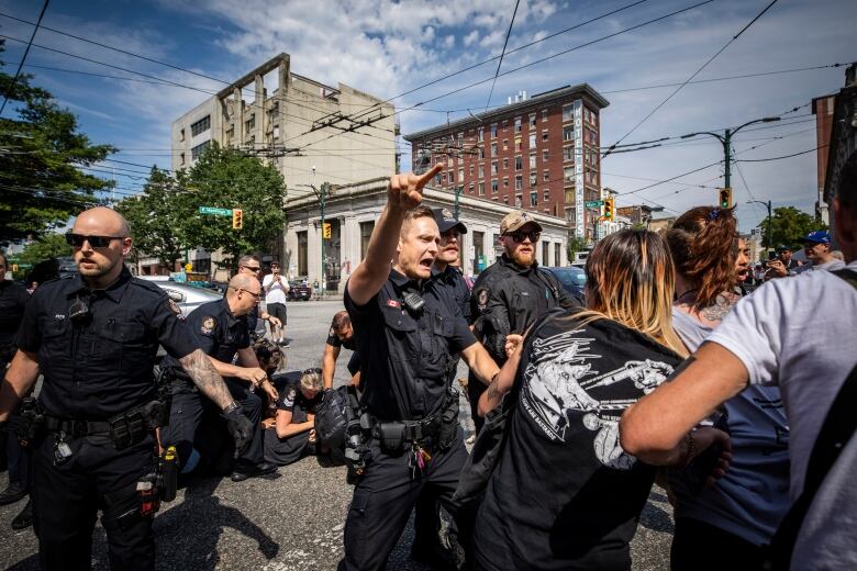 A police officer points authoritatively as others converge on residents on a sidewalk.