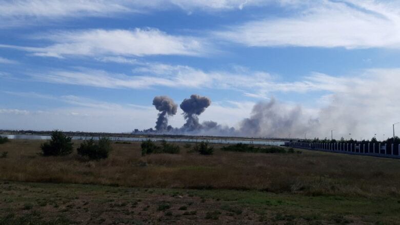 Large clouds of smoke are shown in the distance across a field and body of water.