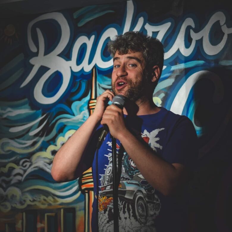 Photo of comedian Brandon Sobel performing on stage at Toronto's Backroom Comedy Club. He is a young white man with short salt-and-pepper hair and a stubbly beard wearing a blue graphic T-shirt. He speaks into a microphone. The backdrop is a graffiti-style mural of the Toronto skyline.