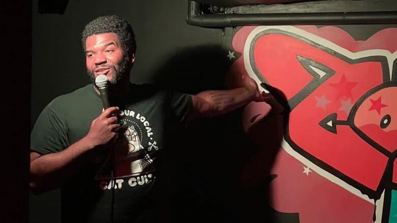 Comedian Danton Lamar onstage at his new Toronto venue, The Comedy Lab. He is a young Black man with short hair and a beard. He wears a black graphic T-shirt and speaks into a microphone. The room is painted black. The wall behind Danton is painted with a graffiti-style design in red.