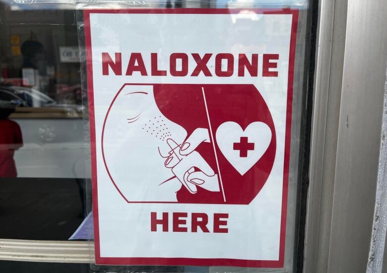 A sign in a window says Naloxone here and has an illustration of a nasal spray. 