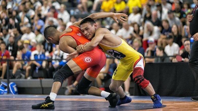 A wrestler with his arms wrapped around his opponent's waist as a large crowd of onlookers watches from the stands. 