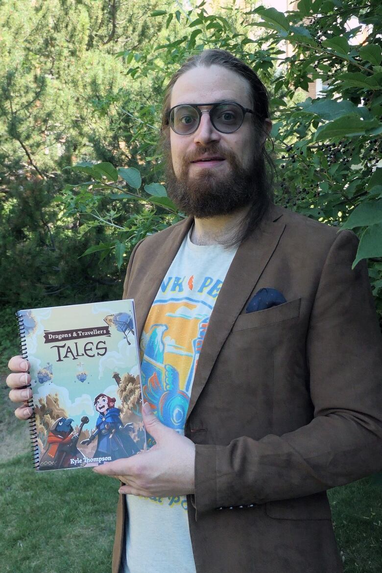 Kyle holds up an illustrated book that has a title 'Dragons' and Travellers' Tales'. He is a white man with a beard wearing round glasses.