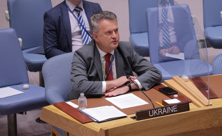 A man in a grey suit sits at a desk with a sign that says 