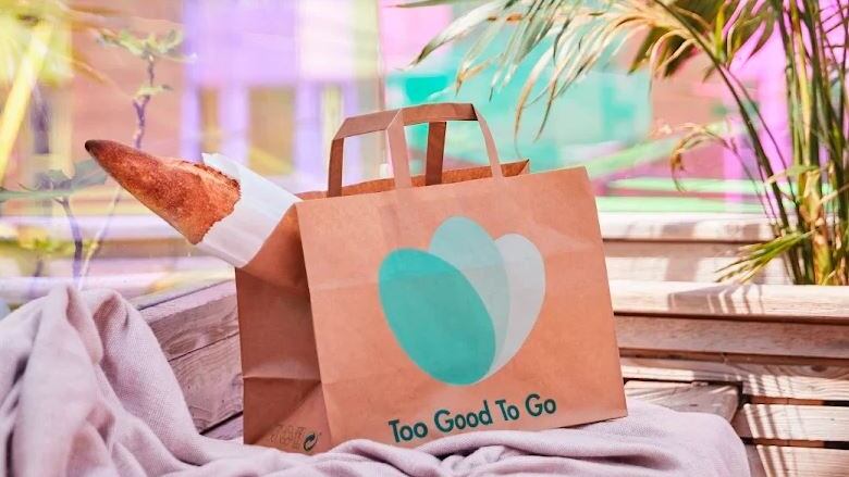 A brown paper bag sits on top of a soft pink tablecloth. On the front of the bag, the Too Good To Go food app is written underneath the app's logo. A baguette sticks out of the left side of the bag.