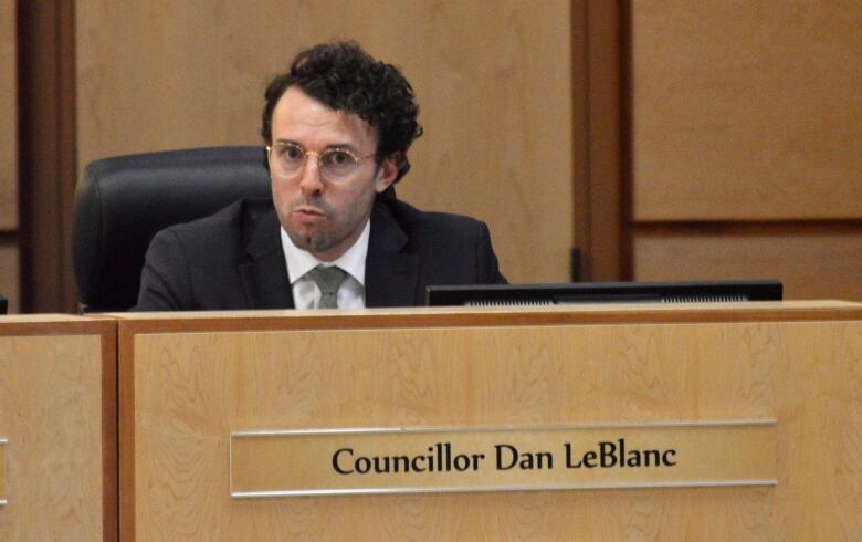 A man with dark, curly hair and glasses in a suit sits at a desk with a sign reading 