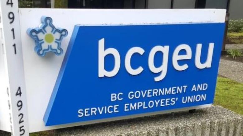 A blue sign with the dogwood flower in the left corner and the words BCGEU and below it B.C. Government and Service Employees' Union rests on a concrete pedestal.