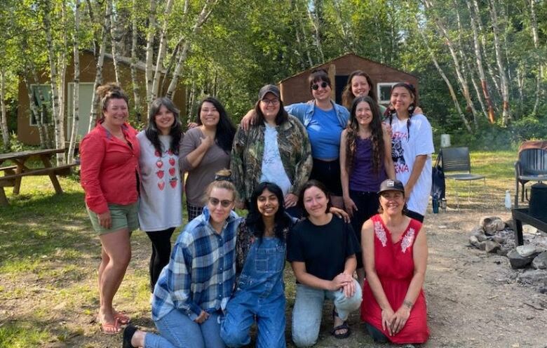 A group of a dozen people -- eight standing and four kneeling in front -- pose in a wooded area, with small buildings, a picnic table and a firepit in the background.