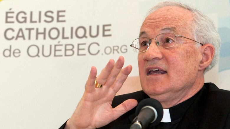 Photo of a cardinal dressed in black and speaking in front of a microphone.