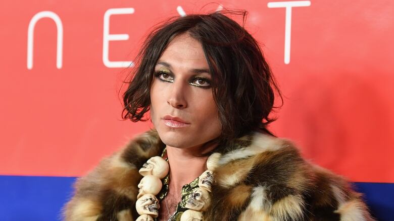 A person with long hair and silver eyeliner poses in front of a photo wall. They are wearing a thick fur coat and a necklace made of fake skulls.