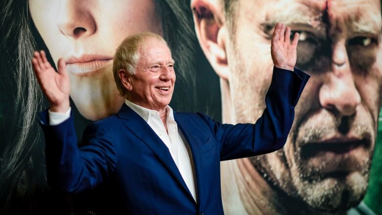 An elderly man in a suit stands smiling in front of a poster depicting the face of a woman and scowling man. 