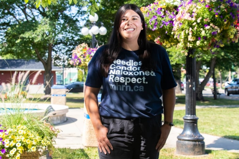 A woman wearing a Manitoba Harm Reduction Network T-shirt stands in a park on a sunny day.