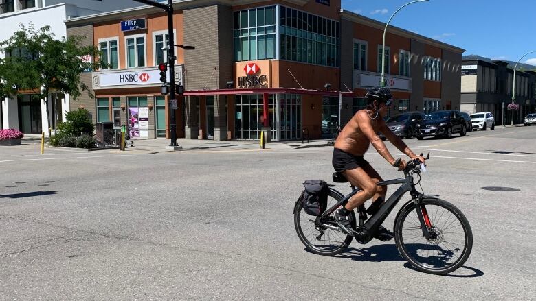 A man without a shirt rides a bike down an empty main road.