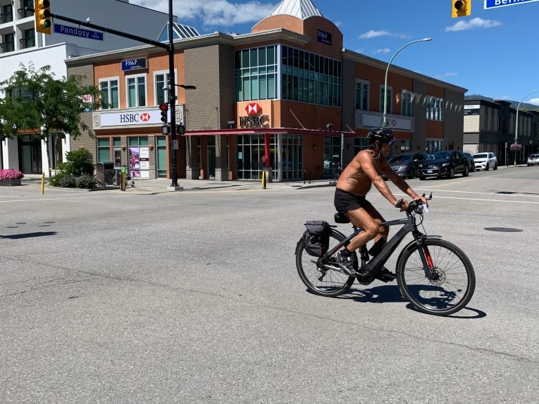 A man without a shirt rides a bike down an empty main road.