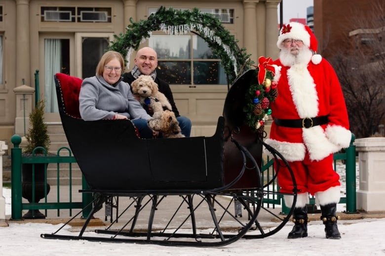 A man, woman and two dogs sit in a Christmas sleigh. A man dressed as Santa stands next to the sleigh.