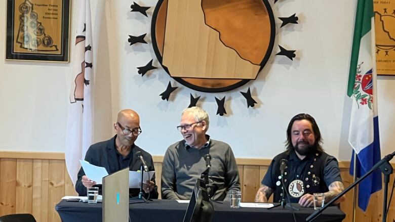 Three men sit at a table with microphones, smiling at one another.