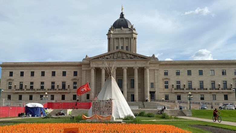 A white teepee sits in front of the Manitoba legislature building with a sea of small, orange flags on the lawn in the foreground.