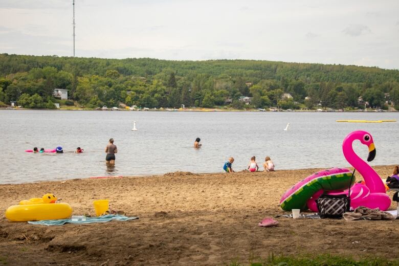 A bright pink plastic floating flamingo is seen on the beach foreground, as several people play in the sand and the water in the background.