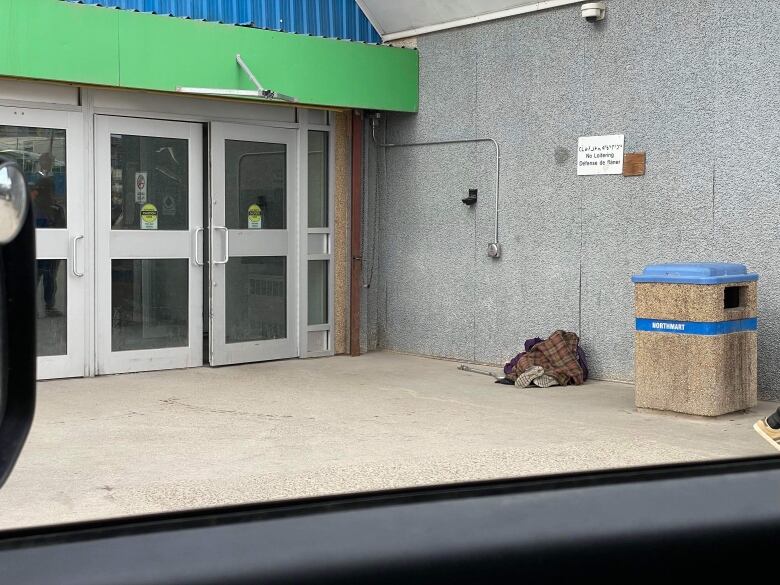 A crumpled body next to a cane is wedged against a stucco wall behind a garbage can in front of two large glass doors.