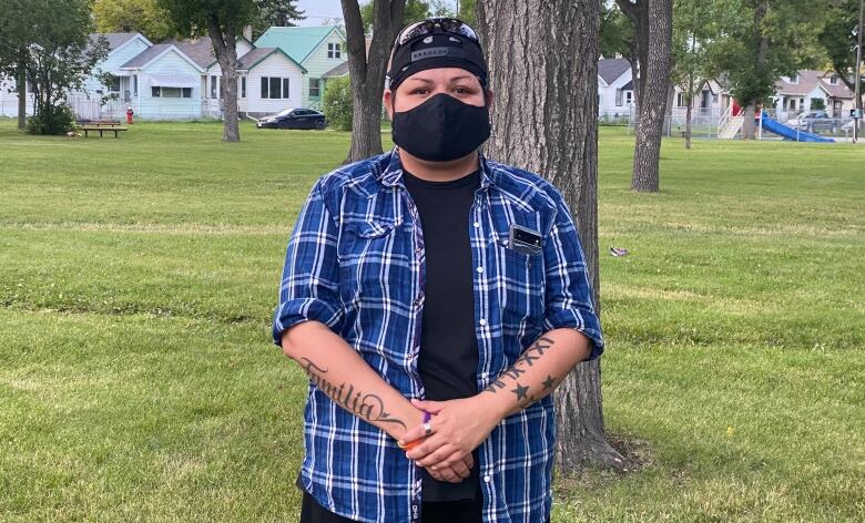 A man wearing a black face mask and a blue checkered shirt stands in a park.