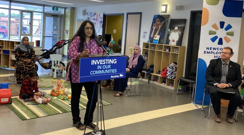 A woman with long black hair wearing a red and white tunic stands behind a podium in a child care facility. The podium has a sign hanging off of it that says 