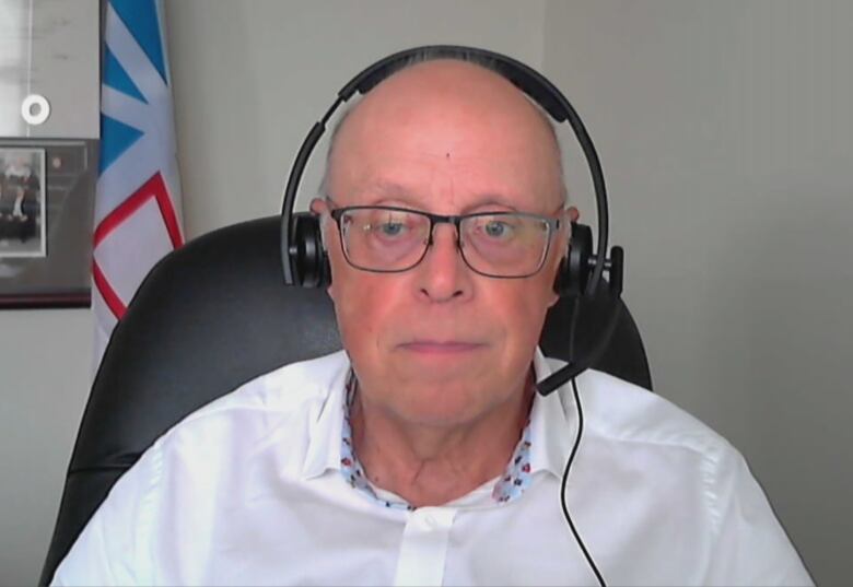 A person wearing a headset sits at a desk, facing the camera. Behind him a Newfoundland and Labrador flag can be seen.