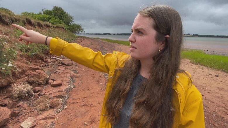 Erin Nelson pointing to coastal erosion with hanging vegetation and the sea in the background.