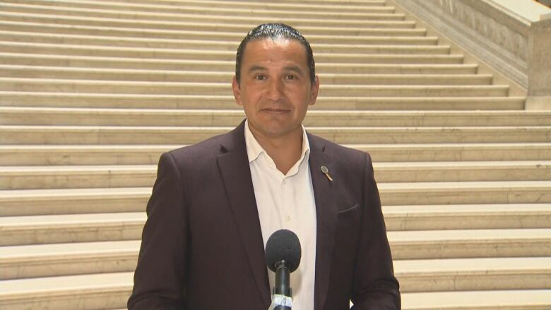 A man wearing a dark suit jacket and a white shirt stands behind a microphone in front of a white stone staircase.