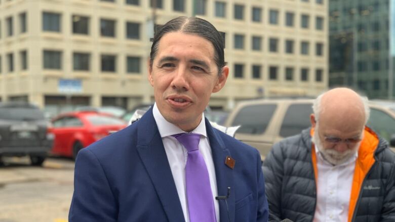 A man in a suit jacket and tie stands in front of microphones with another man behind him. Behind them a number of cars are parked in an outdoor surface lot.