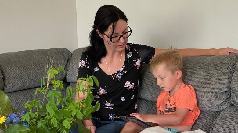 A woman sits with a young boy on a couch. The woman looks down at the boy as he navigates an electronic tablet.