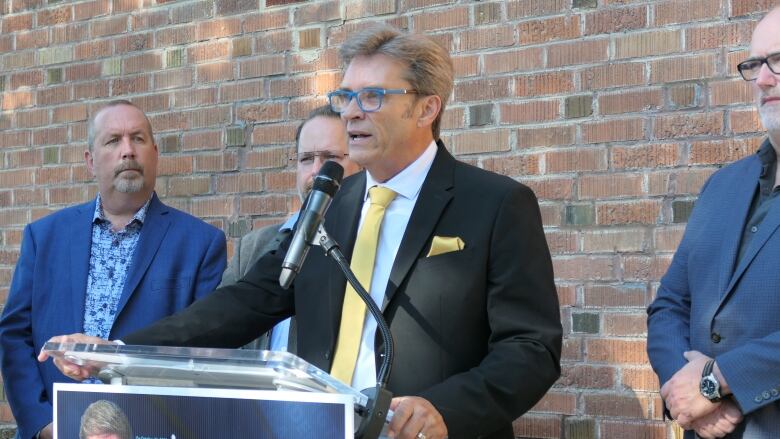 A man wearing a black suit, yellow tie, white shirt and blue-framed glasses speaks at a podium outside. Three men, also wearing suits, stand around him. They are standing in front of a brick wall. 