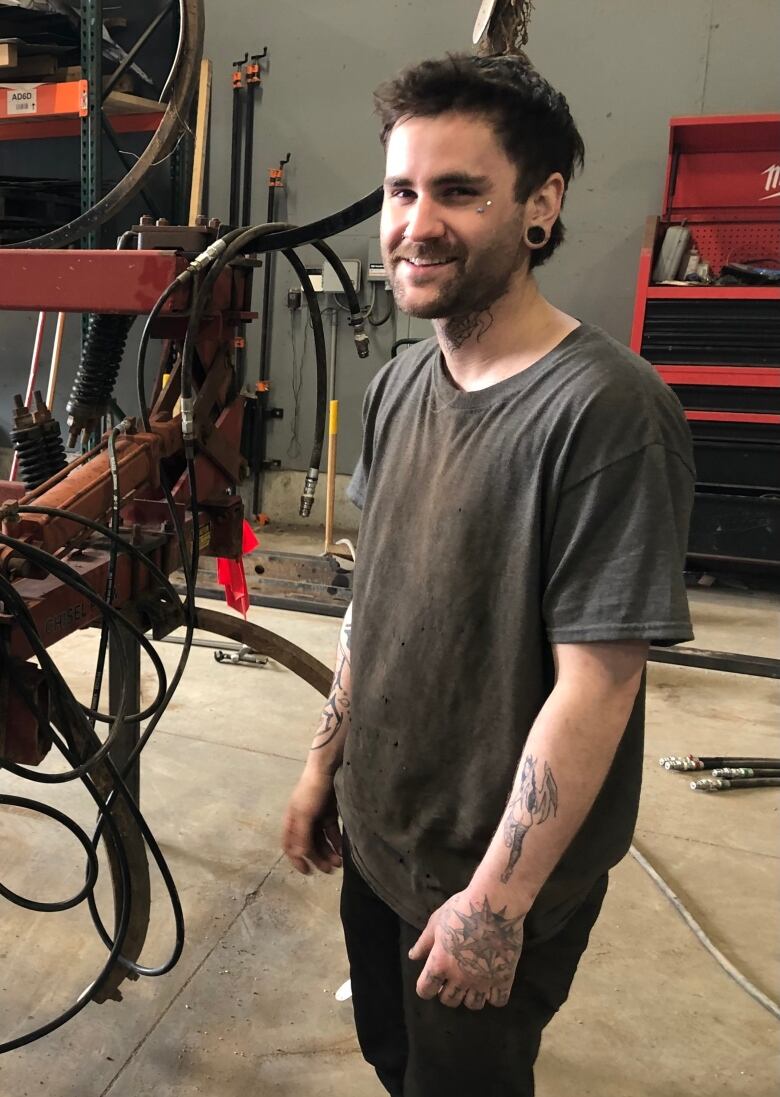 A bearded young man, wearing large stud earrings, a faded grey t-shirt and black pants, with tattoos on his arms stands smiling at the camera, surrounded by machinery in a workshop.