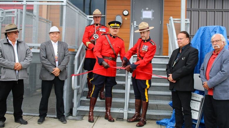 Three uniformed RCMP officers, in red serges, stand outside a building, flanked by four men. Two of the officers hold a red ribbon in front of the building's stair, and one holds a pair of scissor, poised to cut the ribbon.