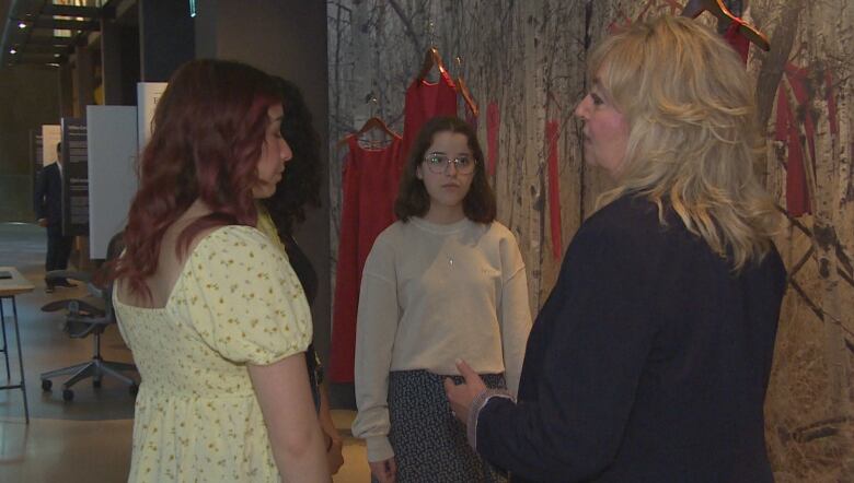 A woman in a dark-coloured jacket stands talking to another woman in a yellow dress and a young woman wearing glasses. They stand in front of a museum display showing red dresses.