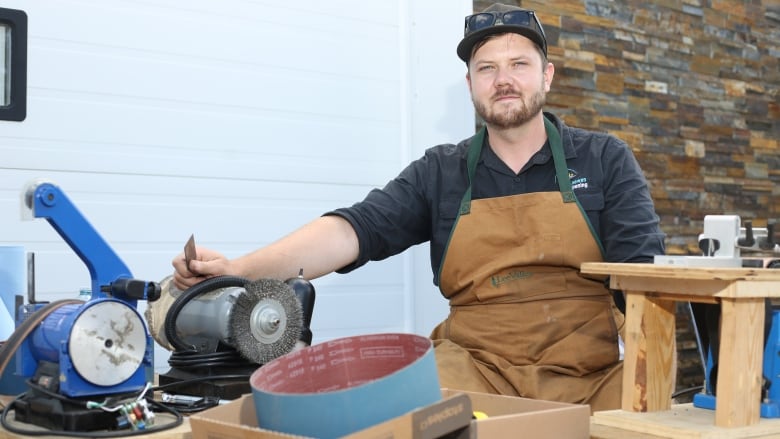 A man in beige overall and a baseball cap sits with his knife sharpening tools.