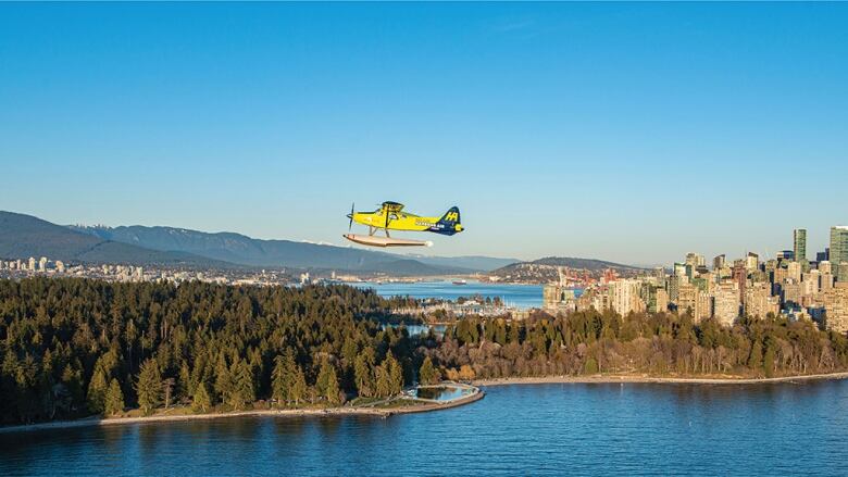 A yellow seaplane flies over a body of water with the Vancouver skyline visible in the background.