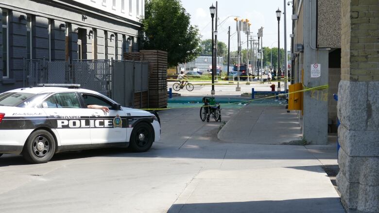 A Winnipeg police vehicle is parked in a lane between two buildings, which is blocked off by police tape. Inside the tape is an empty wheelchair.