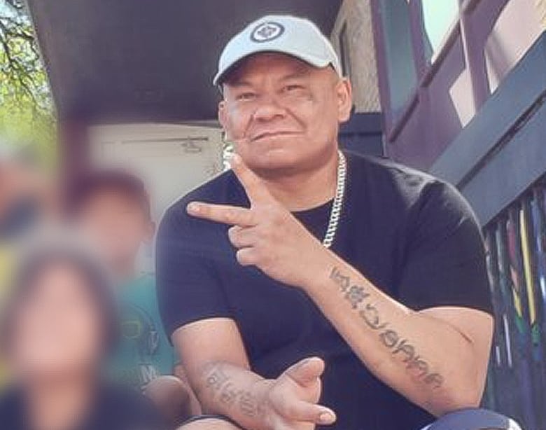 An Indigenous man with a Winnipeg Jets hat on makes the peace symbol towards the camera. He has tattoos all along his arms.