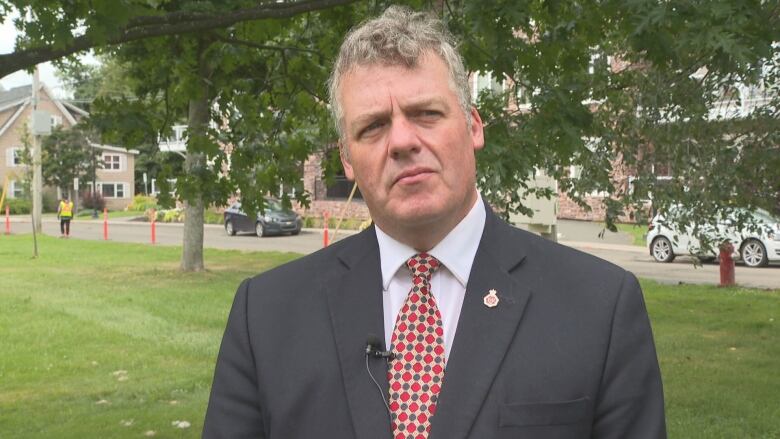 A man in a navy blazer and red tie looks to camera, with a park in background. 