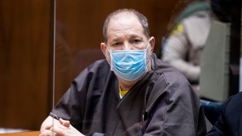 A man wearing a prison uniform and medical mask sits behind glass at a court table. 