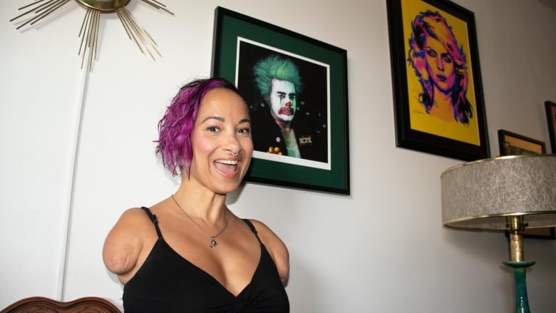 A grinning woman with bright purple hair in a black dress sits in front of a painting of a punk rock singer. The woman does not have arms.