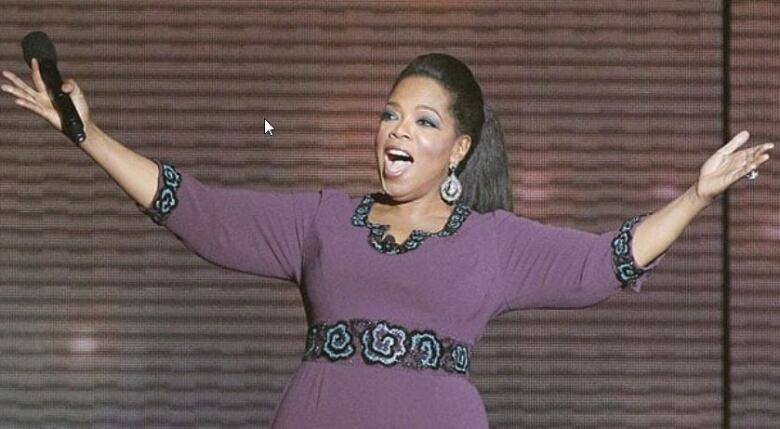 A Black TV show host spreads her arms wide as she greets an audience.