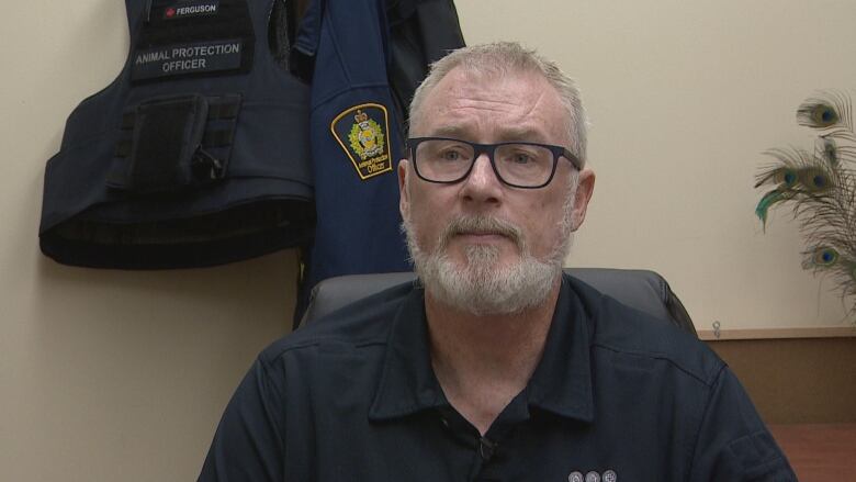 A bearded man with glasses sits in an office, with a uniform jacket and a vest reading 