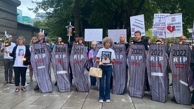 A person holds up a picture of a dead loved one at a protest. They are surrounded by multiple cardboard coffins, with years listed and the words 'RIP'.