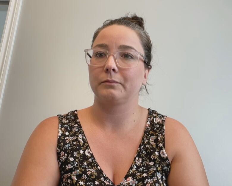 A chest-up shot of a person wearing glasses sitting in front of a blank wall looks into the camera.