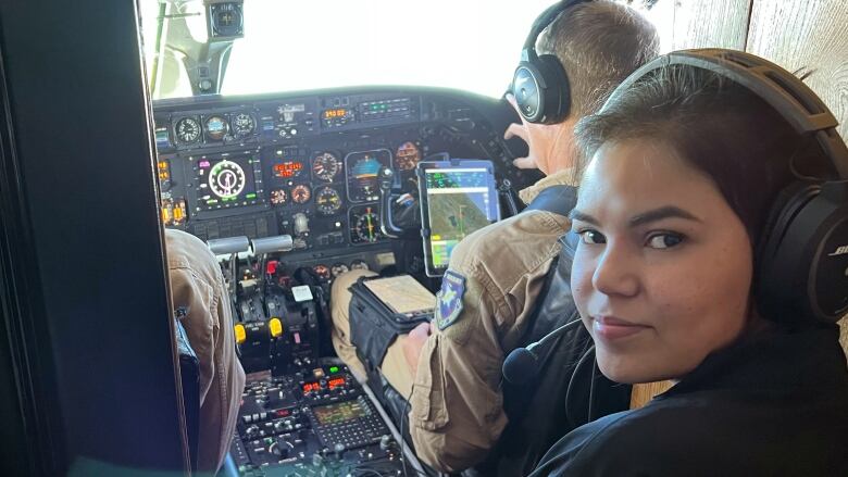 Young woman in headset looks at camera, with 2 pilots in cockpit in background.