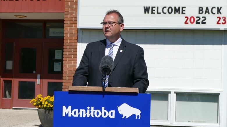 A man in glasses speaks at a podium that says 'Manitoba' on it. Behind him, a marquee has a welcome back message for students returning to classes on it.