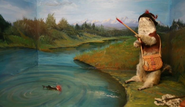 A gopher dressed as a fisherman is depicted as hooking a fish in a quiet pond in a diorama.