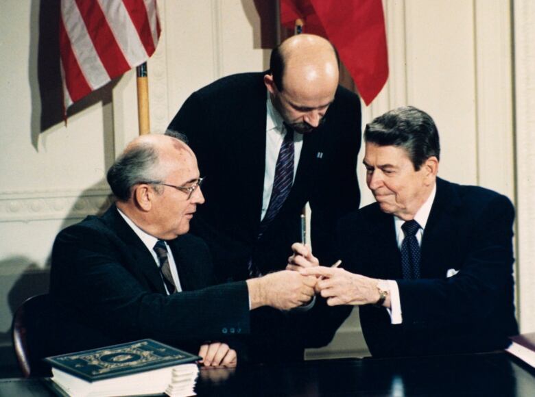 Three men in suits crowded around a table. One is pointing to a book while holding a handful of pens.